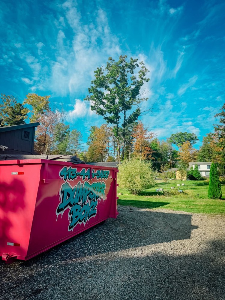The pink Dumpster Boyz dumpster on a gravel driveway beside a green lawn, with a tall maple and a bright blue sky.